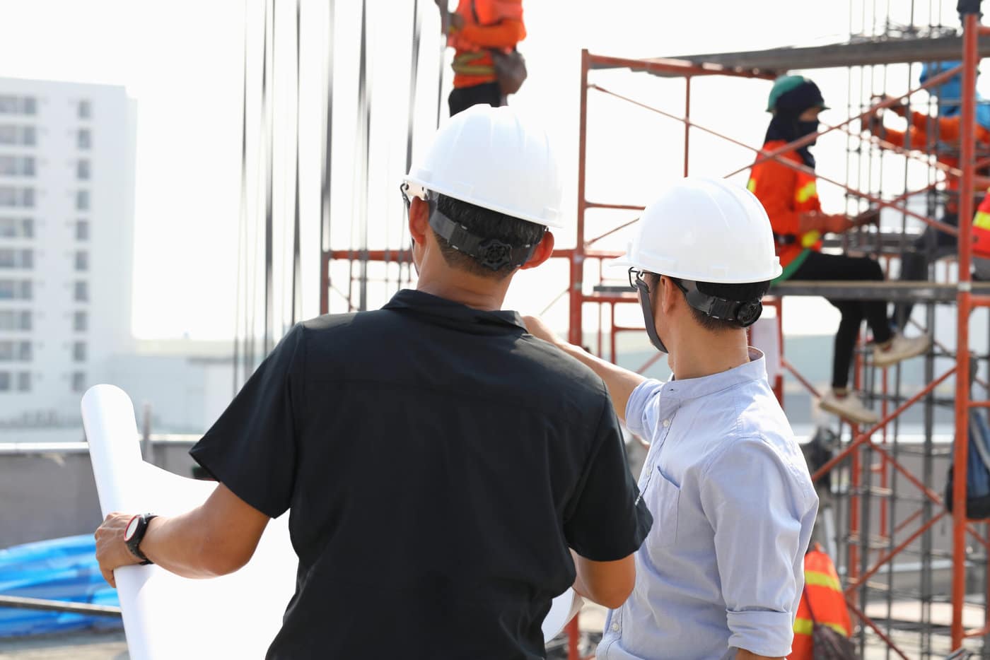 Two Engineers In Hard Hats, One Holding Blueprints, Observing Construction Workers On A Scaffold In An Urban Setting.