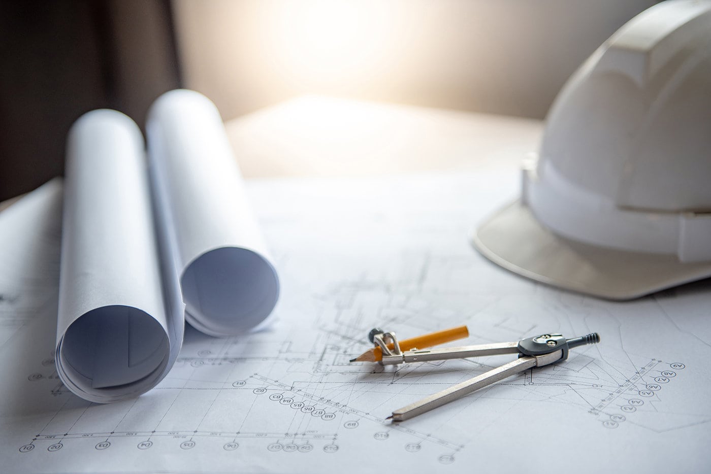 Blueprints, A Compass, And A White Hard Hat On A Table, Illuminated By Sunlight, Suggesting An Architectural Or Engineering Workspace.
